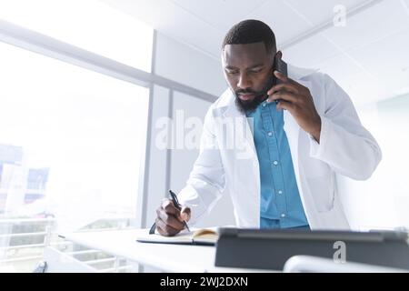 African american male doctor talking on smartphone and taking notes in hospital room, copy space Stock Photo