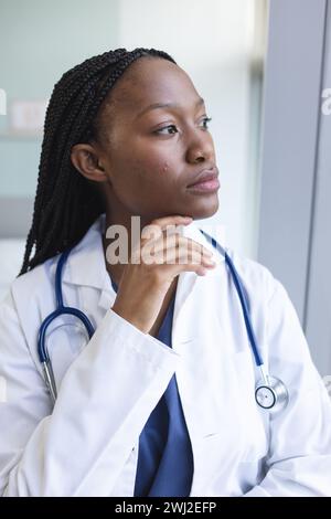 Thoughtful african american female doctor wearing lab coat looking through window in hospital room Stock Photo