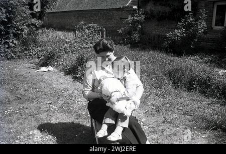 1960s, historical, motherhood, summertime and outside in a country garden of an old farmhouse, an older mother sitting on a chair on the grass, with her young son and new baby, England UK. Stock Photo