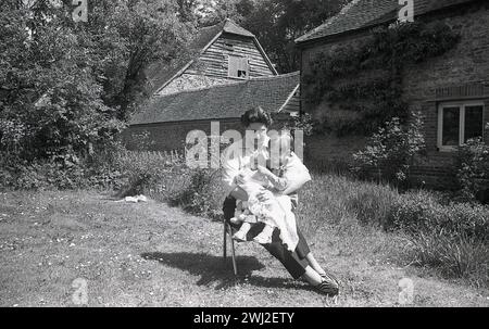 1960s, historical, motherhood, summertime and outside in a country garden of an old farmhouse, an older mother sitting on a chair on the grass, with her young son and new baby, England UK. Stock Photo