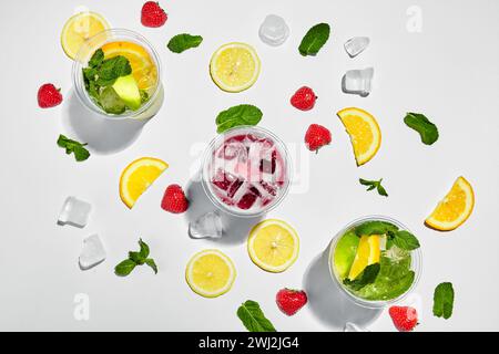 Three glasses with lemonades and ice cubes, lemons, strawberries and mint leaves on white background. View from above Stock Photo