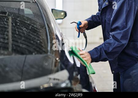 cropped view of dedicated professional worker in uniform holding hose and cleaning car with rag Stock Photo