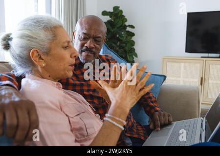 Diverse senior couple sitting on sofa, embracing and using laptop in sunny living room Stock Photo