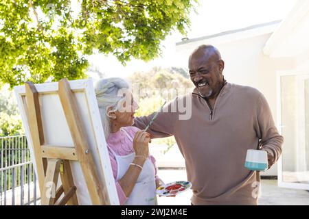 Happy diverse senior couple embracing and looking at painting on sunny terrace Stock Photo