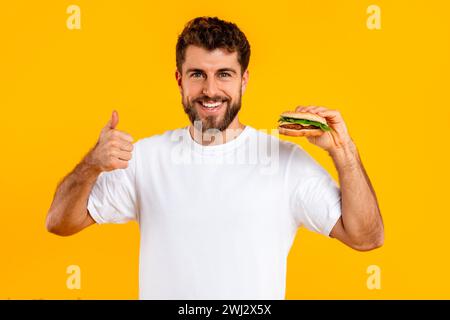 cheerful guy holding cheeseburger gesturing thumbs up on yellow background Stock Photo