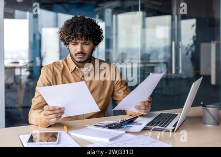 Worried young Indian male businessman working in office with laptop and documents, holding papers and looking disappointed. Stock Photo