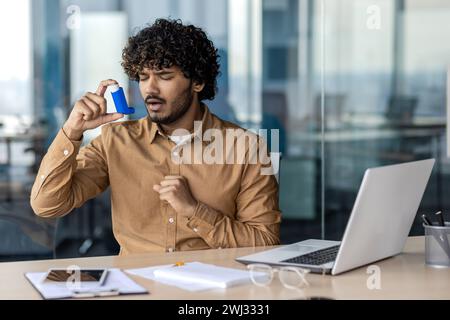 Young Indian man suffering from asthma at workplace, sitting at desk in office and using inhaler. Stock Photo