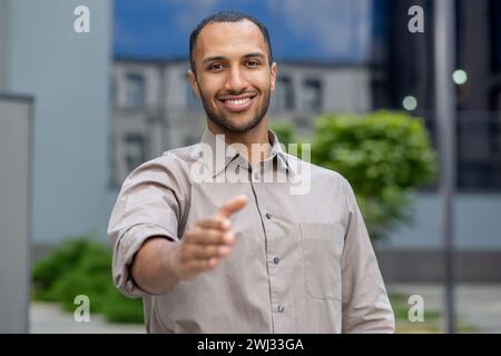 Portrait of a successful and smiling young businessman, hispanic man standing outside an office building and extending his hand to the camera in greeting. Stock Photo