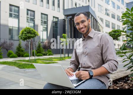 Portrait of young Muslim male student, freelancer sitting outside campus and office center, working and studying on laptop. He looks at the camera with a smile. Stock Photo