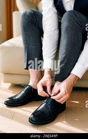 Groom ties his black leather brogues while sitting on the sofa. Cropped. Faceless Stock Photo