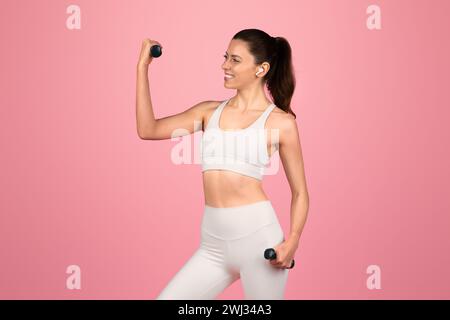 A motivated young woman in white athletic wear confidently flexes her biceps with dumbbells Stock Photo