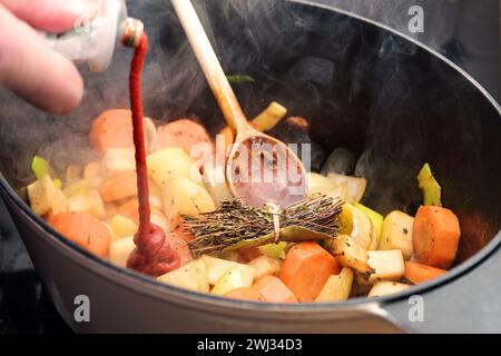 Tomato paste is poured into a casserole pot with steaming roasted vegetables like carrot, onion, celery, leek and a herb bouquet Stock Photo