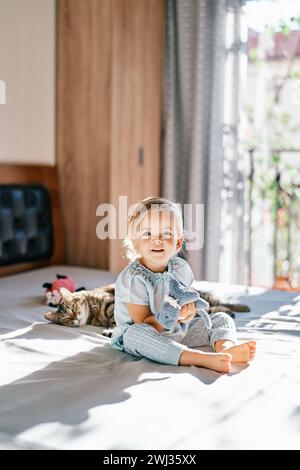 Little smiling girl with a soft toy in her hand sits on the bed next to a sleeping tabby cat Stock Photo
