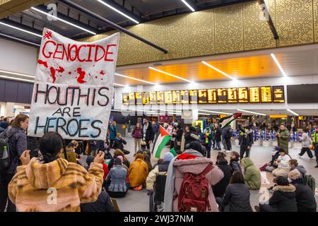 Leeds, UK. 12 FEB, 2024. Protestor holds a 'Genocide/ Houthis are heroes' sign infront of gathered pro palestine demonstrators in front of the main signage boards for Leeds railway station. Credit Milo Chandler/Alamy Live News Stock Photo
