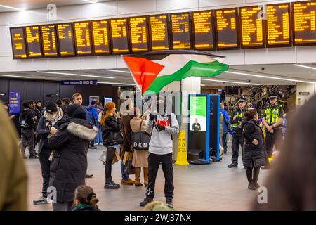 Leeds, UK. 12 FEB, 2024. A masked pro palestine protestor waves palestinian flag infront within the main throughfare for Leeds train station. Credit Milo Chandler/Alamy Live News Stock Photo