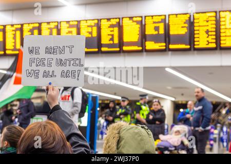 Leeds, UK. 12 FEB, 2024. Protestor holds a sign that reads 'we aren't free until we are all are!' infront of signage and police officers at Leeds Railway station. Credit Milo Chandler/Alamy Live News Stock Photo