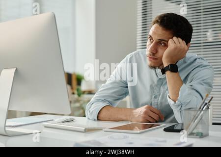 Young bored businessman sitting by workplace in front of desktop computer and looking at screen while waiting for page uploading Stock Photo