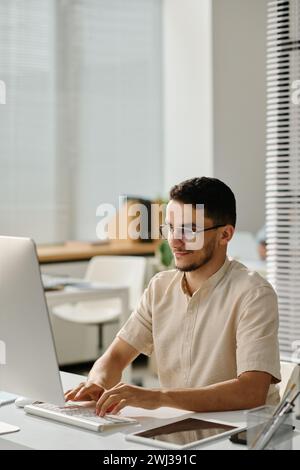 Young Muslim male white collar worker typing on computer keyboard and looking at screen while entering data in electronic document Stock Photo