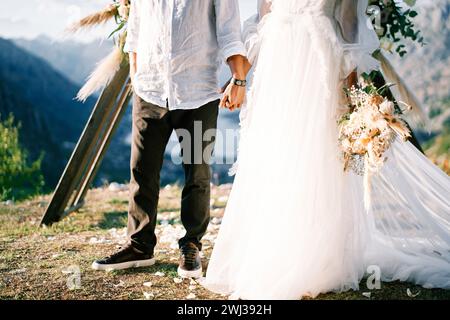 Bride and groom stand holding hands at the wedding arch on a mountain. Cropped. Faceless Stock Photo