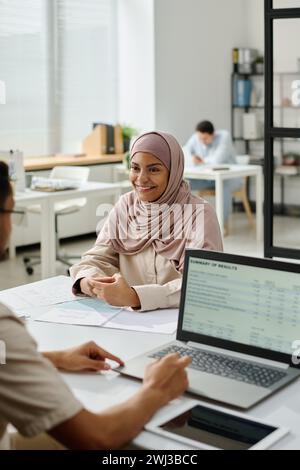Young smiling Muslim female economist in hijab looking at male colleague using laptop while analyzing financial data on screen Stock Photo