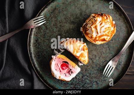 Mini Baked Alaska Desserts with Forks on a Dark Plate: Whole and halved individual servings of bombe Alaska ice cream, cake, and meringue desserts Stock Photo