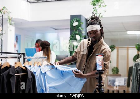 Social media blogger recording video with shirts review for brand promotion in shopping mall. African american marketing specialist speaking about apparel fabric quality on smartphone camera Stock Photo
