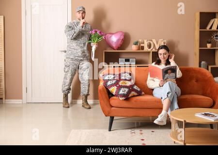 Man in military uniform with bouquet of flowers sneaking to surprise his wife at home. Valentine's Day celebration Stock Photo