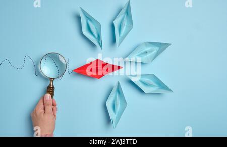 A woman's hand holds a magnifying glass over paper boats, a concept for researching the uniqueness of individuals Stock Photo