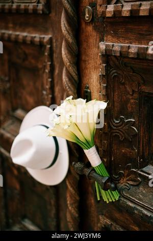 Bridal bouquet and a white hat hang on the metal handle of an antique wooden door Stock Photo