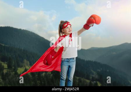 Superhero, motivation and power. Cute girl in cape and boxing gloves on high top in mountains Stock Photo