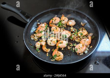 Shrimps with tails are roasted in a frying pan with onions, garlic and herbs on a black stove top, cooking a seafood meal, copy Stock Photo