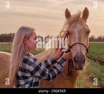 Young blonde girl with a beige horse on the ranch on the background of a green field and evening sky Stock Photo