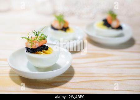Party snacks from boiled eggs with caviar and shrimps on small white plates on a wooden table for a holiday buffet on Christmas, Stock Photo