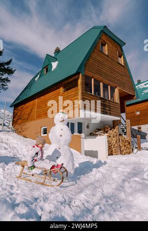Little girl sits on a wooden sled next to a snowman near a wooden cottage. Side view Stock Photo