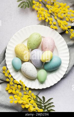 A selection of painted Easter eggs in pastel shades are elegantly displayed on a plate, accented with fresh yellow mimosa blooms. Stock Photo