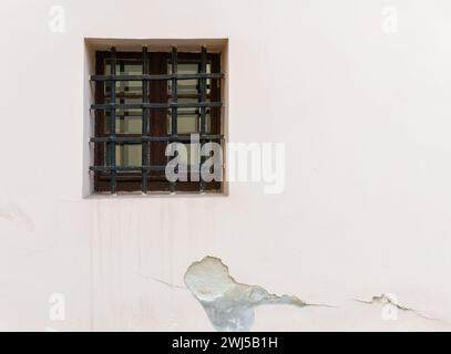 Light beige plaster wall with cracked and chipped plaster and a square old window with wrought iron bars Stock Photo