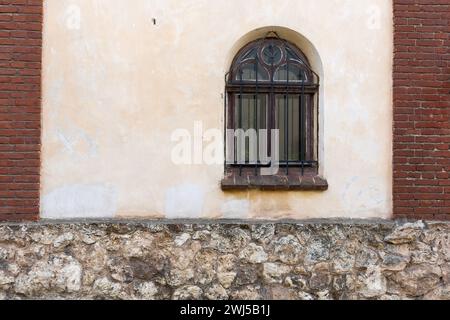 Wall of an old house with a window with a wrought-iron lattice with the upper part in the form of a semicircular arch Stock Photo