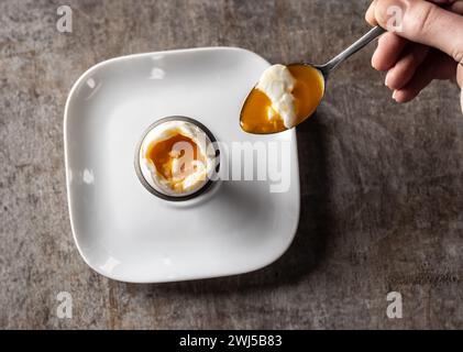 A woman's hand with a spoon scoops a soft-boiled egg from the shell - Top of view. Stock Photo