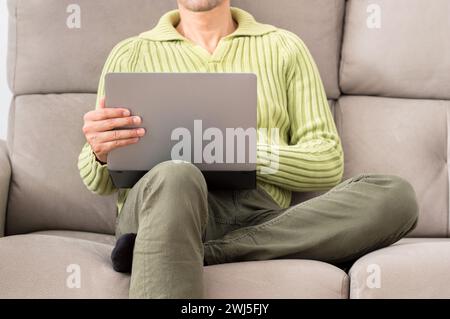 Front view of a legs of a man looking a laptop sitting on a sofa in the living room in a house interior in winter Stock Photo