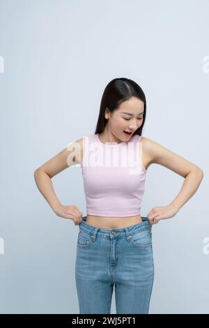Young smiling happy woman show loose pants on waist after weightloss isolated on white background. Stock Photo