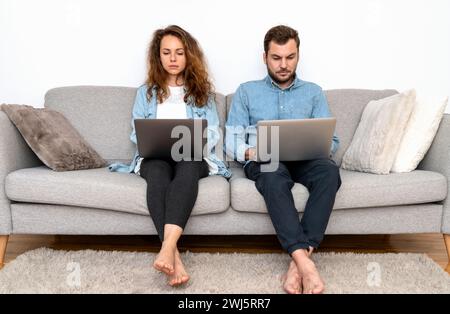 serious and focused man and woman sit on sofa next ti each other and using laptops. Stock Photo