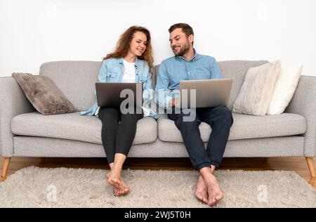 Woman and man sit on couch using their laptops and smiling. Online shopping. Stock Photo
