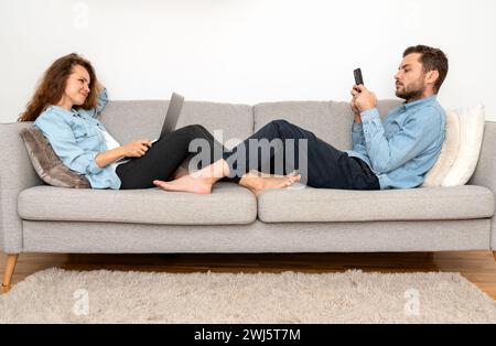 Young adult couple relaxing on sofa at home and using their personal digital devices. Stock Photo
