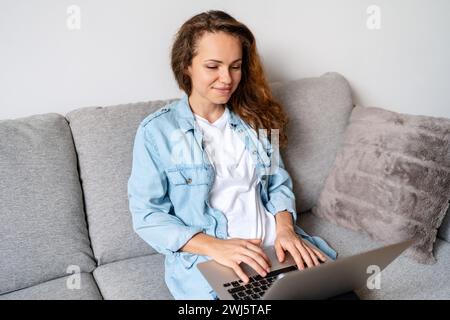 Brunette woman in denim shirt sitting on sofa at home and using laptop. Online shopping. Working from home. Stock Photo