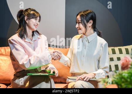 Two happy young asian businesswomen shaking hands watching each other, sitting on the sofa in the office. Stock Photo
