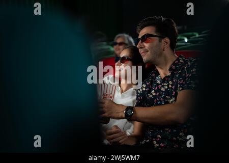 In a cinema, A young couple pair wearing 3D glasses watches movies and eats popcorn. Stock Photo