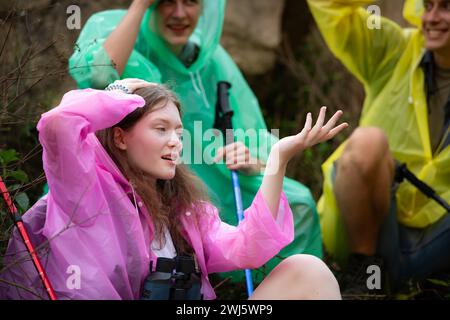 Group of friends in raincoat with backpacks hiking in the forest, Sit and shelter from the rain Stock Photo