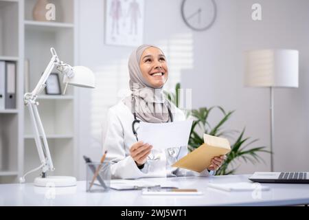 Smiling female Muslim doctor in a hijab and white coat holding papers in a modern clinic office, showcasing professional healthcare. Stock Photo