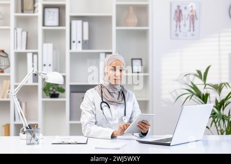 Professional Muslim female doctor in a hijab and white coat working at her office, using a tablet, with laptop and stethoscope on desk. Stock Photo