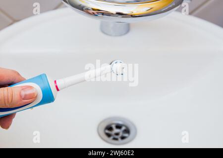 Hand holding electric toothbrush with toothpaste in bathroom near ceramic faucet. Daily dental hygiene routine Stock Photo